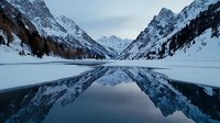 Majestic snow-capped mountain peaks perfectly mirrored in still alpine lake waters during winter twilight, creating symmetrical reflection against moody sky.