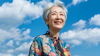 Senior Asian woman with short gray hair wearing traditional floral qipao dress smiling against blue sky with white clouds, expressing joy and confidence in outdoor portrait.