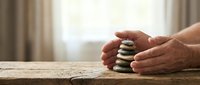 Hands protecting balanced zen stones on wooden table symbolizing mindfulness, meditation, wellness and spiritual harmony in peaceful home setting.