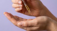 Hands applying golden serum oil drops from pipette dropper for skincare routine and beauty treatment on purple background.