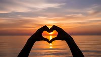 Hands forming heart shape silhouette against stunning ocean sunset with golden sky reflections on calm water surface creating romantic atmosphere.