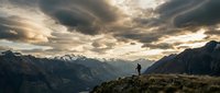 Hiker standing on mountain ridge overlooking vast alpine valley with dramatic cloudy sky and snow-capped peaks, outdoor adventure and wilderness exploration.