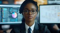 Young African American businesswoman in glasses and suit working late at office with financial data and charts displayed on monitors in background.