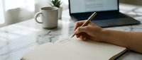 Person writing in notebook at marble desk with coffee cup and laptop in background.