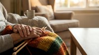 Elderly woman's hands resting on colorful knitted blanket with knitting needles in cozy living room, warm home atmosphere.