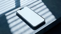 Modern smartphone with blank white screen lying on dark surface with dramatic window blind shadows creating striped light pattern for mockup design.