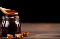 Liquid caramel sauce dripping from wooden spoon into glass jar with caramel candy cubes on rustic wooden table against dark background, sweet food photography.