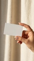 Female hand holding blank white business card mockup with natural sunlight shadows on neutral beige background for branding presentation.