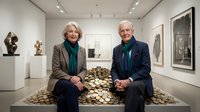 Mature couple, Caucasian, sitting together on golden coins in contemporary art gallery surrounded by sculptures and framed artwork, representing wealth, investment, financial planning, retirement savings.