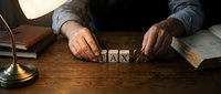 Mature male hands arranging wooden tax blocks on desk with lamp and documents, representing financial planning and accounting concepts for business and tax preparation.