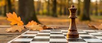 Wooden chess queen piece standing on chessboard with autumn maple leaves in blurred forest background, strategic thinking and decision making concept.
