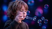 Young blonde boy blowing soap bubbles with concentrated expression against colorful bokeh background. Childhood play and wonder concept for family content.