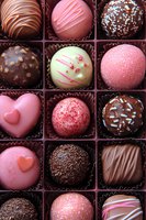 Assorted luxury chocolate truffles and bonbons in pink, white and dark chocolate with decorative toppings arranged in brown paper cups, featuring heart shapes and sprinkles.