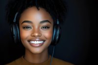 Young African American woman with bright smile wearing headphones against dark background, natural makeup and dreadlocks hairstyle in casual brown sweater.
