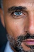 Close-up portrait of middle eastern man with striking blue eyes, dark beard and gentle expression against dark background, showing partial face detail.