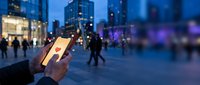 Person using smartphone in busy urban evening scene with blurred city lights and pedestrians walking in background creating bokeh effect atmosphere.