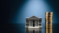 Miniature bank building with classical columns next to stack of golden coins on reflective surface representing financial services and banking concepts.