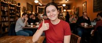 Young woman giving thumbs up gesture while sitting in cozy bookstore cafe with warm lighting and people studying in background.