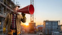 Construction foreman using red megaphone at building site during sunset for team communication and project management in urban development zone.