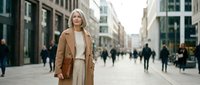 Confident blonde businesswoman in beige coat walking through modern city street during daytime with urban architecture and pedestrians in background.