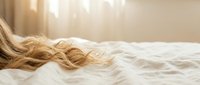 Blonde woman sleeping peacefully in bed with flowing wavy hair spread across white pillows in soft morning sunlight streaming through window.