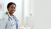 Confident African American female doctor smiling in modern medical office wearing white coat and stethoscope for healthcare and medical consultation.