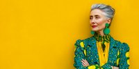 Confident senior woman with gray hair wearing bright green and yellow outfit against vibrant yellow wall, arms crossed, looking sideways.
