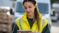 Young Hispanic female warehouse worker in safety vest checking digital tablet while managing logistics operations in outdoor industrial setting.