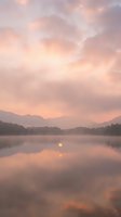 Serene mountain lake at sunrise with soft pink and gold clouds reflecting on calm water, misty forest shoreline, peaceful dawn landscape for nature and travel projects.