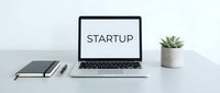 Laptop on a clean desk displaying the word Startup, with a notebook, pen, and small plant, representing modern entrepreneurship and business launch concepts.