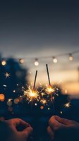 Two lit sparklers held in hands against twilight sky with string lights and bokeh background, celebrating festive occasions and special moments.