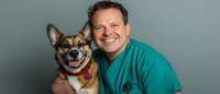 Smiling veterinarian in green scrubs embracing happy Shiba Inu dog during checkup, showcasing positive pet healthcare experience against studio background.