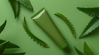 Green cosmetic tube with aloe vera leaves scattered on green background, minimalist skincare product photography for beauty and wellness marketing.