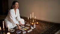 Young Caucasian woman in white robe performing tarot card reading by candlelight on ornate carpet creating mystical spiritual atmosphere for meditation.