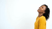 Young Black woman with long braided hair wearing a yellow shirt, laughing joyfully with head tilted back against white background, expressing happiness and positivity for lifestyle and wellness content.