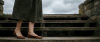 Woman in elegant nude heels and long coat standing on ancient stone steps. Minimalist fashion photography with moody atmospheric lighting for luxury brands.