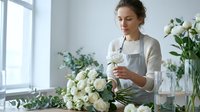 Young woman, Caucasian ethnicity, florist professional arranging white roses and greenery in vase by bright window. Floral design and arrangement work.