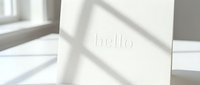 White greeting card with embossed hello text standing in soft natural window light with gentle shadows, minimal elegant design for stationery.