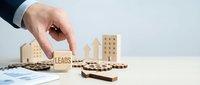 Business professional hand selecting wooden block with leads text among house models representing real estate lead generation and property marketing strategy.