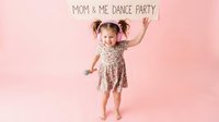 Happy toddler girl with pigtails dancing in floral dress on pink background for mom and me dance party celebration with raised arms showing joy and movement.