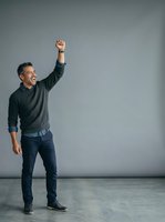 Middle-aged man with dark hair and beard, wearing charcoal sweater and navy jeans, raising fist in victory gesture against neutral gray background for success and achievement concepts.
