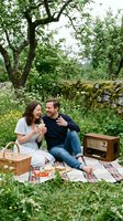 Couple enjoying outdoor picnic in countryside garden with vintage radio, wicker basket, and plaid blanket surrounded by lush green trees and stone wall.