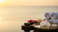 Spa wellness setup with candles, towels and orchid flowers by ocean at sunset for relaxation and massage therapy treatments.