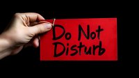 Hand holding bright red door hanger sign with black handwritten text saying do not disturb against dark background, concept of privacy and personal space.