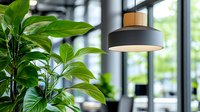Modern pendant lamp with wooden accent hanging in bright office space with lush green tropical plant leaves in foreground, creating contemporary eco-friendly workspace atmosphere.