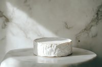 Whole round of soft ripened cheese with white bloomy rind on marble surface in natural light, minimalist food photography composition for gourmet content.