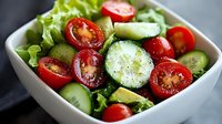 Fresh garden salad with sliced cherry tomatoes, crisp cucumber rounds, and green lettuce leaves in white ceramic bowl, seasoned with black pepper.
