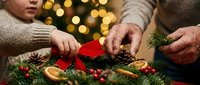 Hands crafting festive holiday wreath with pine cones, dried oranges and red ribbon. Christmas decoration making activity with warm bokeh lights background.