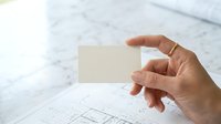 Architect or designer holding blank business card over construction blueprints on marble surface, suitable for professional branding and architectural services marketing.