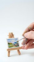 Hand holding paintbrush applying watercolor strokes to miniature landscape artwork on tiny wooden easel. Creative hobby and artistic expression concept for design and illustration projects.